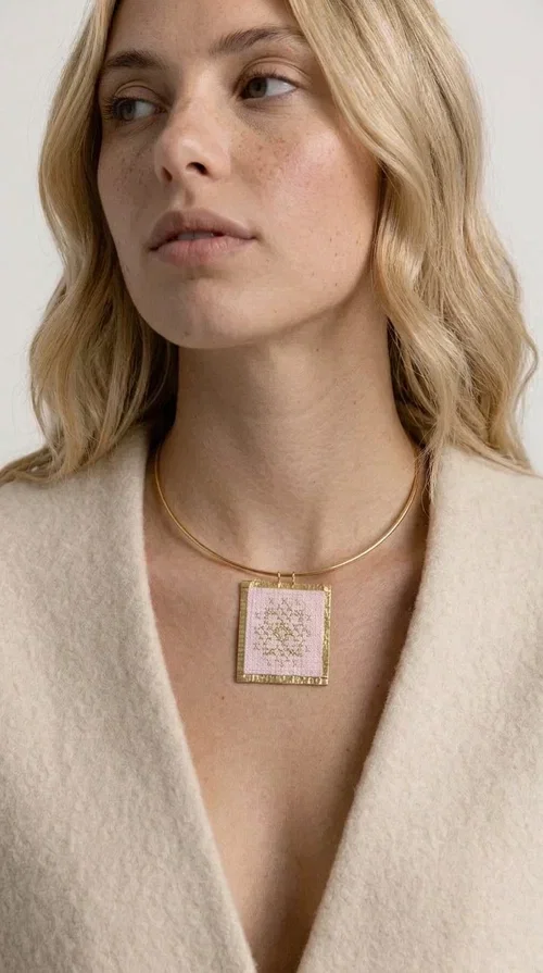 , The statement embroidered necklace in a pink hue is a piece of jewelry that combines the traditional art of embroidery with contemporary design, creating a result that immediately catches the eye. Designed by Aikaterini Chalkiadaki, it carries the signature of Greek handmade craftsmanship and is an ideal choice for those seeking jewelry with character and history.

The necklace features a rectangular embroidered pendant with geometric patterns inspired by folk art and a folklore aesthetic. The pink fabric base combined with golden brass details creates a harmonious contrast that highlights the handmade element. The gold-toned metal omega-style chain gives structure and elegance to the whole, while the pendant is framed by distinctive golden details that emphasize the craftsmanship.

Wear it over a white t-shirt for a casual look or pair it with a blazer for a professional or evening look — just as shown in the photo. It is a piece of jewelry that suits every style and makes an excellent gift idea.
