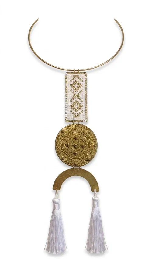, The statement embroidered necklace with geometric patterns in white and gold is a piece of jewelry that never goes unnoticed — it combines folk tradition with a contemporary aesthetic in a way that only handmade artistry can achieve. Designed by Greek Jewelry Designer Aikaterini Chalkiadaki, this necklace is a tribute to Greek craftsmanship and a love for detailed, inspired design.

The necklace is worn on a snug brass base (torque-style choker), from which hangs an embroidered rectangular piece featuring traditional geometric motifs in white and gold. Beneath it, a striking gold brass disc with embossed designs and a semicircular element complete the composition, while two white tassels add movement and boho-chic character. The contrast of white fabric and gold brass creates a result that is both vintage and modern at the same time.

Perfect for summer looks, beach events, outdoor weddings, or everyday boho outfits. It pairs beautifully with simple, solid-colored clothing — just as shown in the photo with the dark green sleeveless dress — letting the necklace speak for itself.