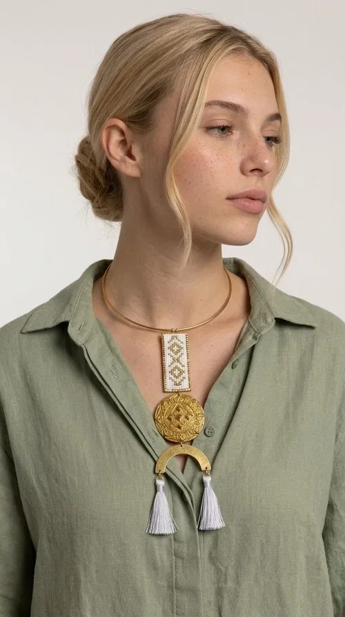 , The statement embroidered necklace with geometric patterns in white and gold is a piece of jewelry that never goes unnoticed — it combines folk tradition with a contemporary aesthetic in a way that only handmade artistry can achieve. Designed by Greek Jewelry Designer Aikaterini Chalkiadaki, this necklace is a tribute to Greek craftsmanship and a love for detailed, inspired design.

The necklace is worn on a snug brass base (torque-style choker), from which hangs an embroidered rectangular piece featuring traditional geometric motifs in white and gold. Beneath it, a striking gold brass disc with embossed designs and a semicircular element complete the composition, while two white tassels add movement and boho-chic character. The contrast of white fabric and gold brass creates a result that is both vintage and modern at the same time.

Perfect for summer looks, beach events, outdoor weddings, or everyday boho outfits. It pairs beautifully with simple, solid-colored clothing — just as shown in the photo with the dark green sleeveless dress — letting the necklace speak for itself.