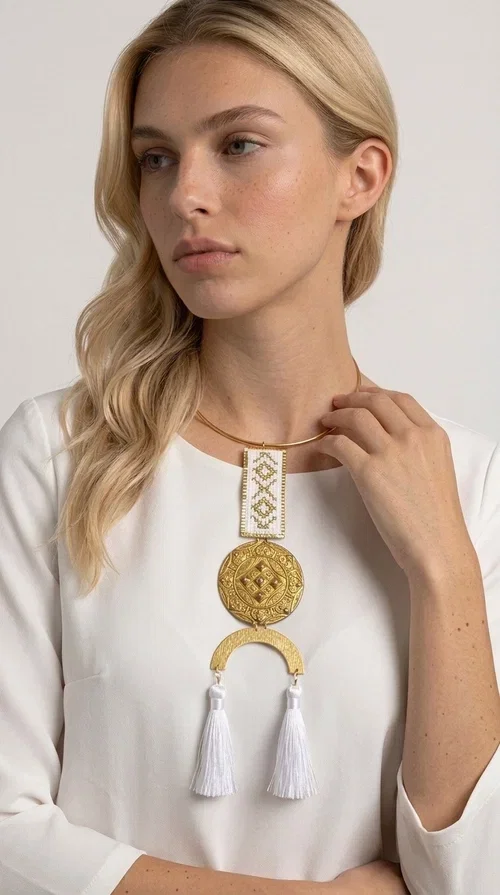 , The statement embroidered necklace with geometric patterns in white and gold is a piece of jewelry that never goes unnoticed — it combines folk tradition with a contemporary aesthetic in a way that only handmade artistry can achieve. Designed by Greek Jewelry Designer Aikaterini Chalkiadaki, this necklace is a tribute to Greek craftsmanship and a love for detailed, inspired design.

The necklace is worn on a snug brass base (torque-style choker), from which hangs an embroidered rectangular piece featuring traditional geometric motifs in white and gold. Beneath it, a striking gold brass disc with embossed designs and a semicircular element complete the composition, while two white tassels add movement and boho-chic character. The contrast of white fabric and gold brass creates a result that is both vintage and modern at the same time.

Perfect for summer looks, beach events, outdoor weddings, or everyday boho outfits. It pairs beautifully with simple, solid-colored clothing — just as shown in the photo with the dark green sleeveless dress — letting the necklace speak for itself.