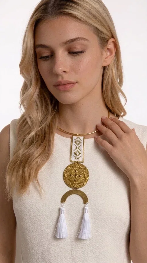 , The statement embroidered necklace with geometric patterns in white and gold is a piece of jewelry that never goes unnoticed — it combines folk tradition with a contemporary aesthetic in a way that only handmade artistry can achieve. Designed by Greek Jewelry Designer Aikaterini Chalkiadaki, this necklace is a tribute to Greek craftsmanship and a love for detailed, inspired design.

The necklace is worn on a snug brass base (torque-style choker), from which hangs an embroidered rectangular piece featuring traditional geometric motifs in white and gold. Beneath it, a striking gold brass disc with embossed designs and a semicircular element complete the composition, while two white tassels add movement and boho-chic character. The contrast of white fabric and gold brass creates a result that is both vintage and modern at the same time.

Perfect for summer looks, beach events, outdoor weddings, or everyday boho outfits. It pairs beautifully with simple, solid-colored clothing — just as shown in the photo with the dark green sleeveless dress — letting the necklace speak for itself.