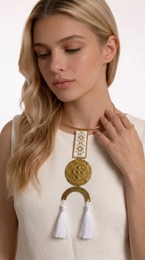 , The statement embroidered necklace with geometric patterns in white and gold is a piece of jewelry that never goes unnoticed — it combines folk tradition with a contemporary aesthetic in a way that only handmade artistry can achieve. Designed by Greek Jewelry Designer Aikaterini Chalkiadaki, this necklace is a tribute to Greek craftsmanship and a love for detailed, inspired design.

The necklace is worn on a snug brass base (torque-style choker), from which hangs an embroidered rectangular piece featuring traditional geometric motifs in white and gold. Beneath it, a striking gold brass disc with embossed designs and a semicircular element complete the composition, while two white tassels add movement and boho-chic character. The contrast of white fabric and gold brass creates a result that is both vintage and modern at the same time.

Perfect for summer looks, beach events, outdoor weddings, or everyday boho outfits. It pairs beautifully with simple, solid-colored clothing — just as shown in the photo with the dark green sleeveless dress — letting the necklace speak for itself.