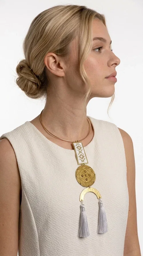 , The statement embroidered necklace with geometric patterns in white and gold is a piece of jewelry that never goes unnoticed — it combines folk tradition with a contemporary aesthetic in a way that only handmade artistry can achieve. Designed by Greek Jewelry Designer Aikaterini Chalkiadaki, this necklace is a tribute to Greek craftsmanship and a love for detailed, inspired design.

The necklace is worn on a snug brass base (torque-style choker), from which hangs an embroidered rectangular piece featuring traditional geometric motifs in white and gold. Beneath it, a striking gold brass disc with embossed designs and a semicircular element complete the composition, while two white tassels add movement and boho-chic character. The contrast of white fabric and gold brass creates a result that is both vintage and modern at the same time.

Perfect for summer looks, beach events, outdoor weddings, or everyday boho outfits. It pairs beautifully with simple, solid-colored clothing — just as shown in the photo with the dark green sleeveless dress — letting the necklace speak for itself.
