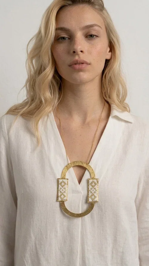 , An embroidered necklace that blends tradition with modern design — the Statement necklace KOL-4745 is the ideal choice for every woman who wants to express herself with style and authenticity.

Designed by Greek Jewelry Designer Aikaterini Chalkiadaki, this striking long necklace features a handmade oval brass element in a gold tone, framed by two rectangular embroidered plaques made of white fabric with geometric patterns inspired by Greek folk tradition. The gold-tone chain adds an elegant, modern touch, while the embroidered motifs bring a warm, handcrafted feel.

Perfect for pairing with simple outfits in neutral shades — such as khaki, white or beige — it turns any look into a true statement. Suitable for both everyday wear and special evening occasions.