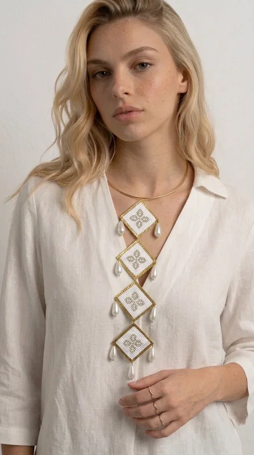 , An embroidered statement necklace that instantly catches the eye — featuring geometric patterns inspired by Greek tradition and an aesthetic that speaks to both the past and the present.

Designed by Greek Jewelry Designer Aikaterini Chalkiadaki, this statement necklace combines white embroidered fabric with gold brass and white pearls in a composition reminiscent of folklore art with a contemporary touch. Four diamond-shaped embroidered elements are stacked vertically, one beneath the other, with teardrop pearls gracefully hanging from each corner. The piece is completed with a delicate gold rigid choker, giving it an airy yet striking presence.

Ideal for women who love long, eye-catching necklaces with a distinctive identity. It pairs perfectly with simple, solid-colored outfits — just as shown in the photo with the pink sleeveless top — or with boho looks for festivals and summer outings. It can also be combined with matching necklace and earring sets for a complete look.