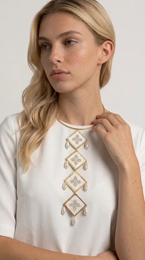 , An embroidered statement necklace that instantly catches the eye — featuring geometric patterns inspired by Greek tradition and an aesthetic that speaks to both the past and the present.

Designed by Greek Jewelry Designer Aikaterini Chalkiadaki, this statement necklace combines white embroidered fabric with gold brass and white pearls in a composition reminiscent of folklore art with a contemporary touch. Four diamond-shaped embroidered elements are stacked vertically, one beneath the other, with teardrop pearls gracefully hanging from each corner. The piece is completed with a delicate gold rigid choker, giving it an airy yet striking presence.

Ideal for women who love long, eye-catching necklaces with a distinctive identity. It pairs perfectly with simple, solid-colored outfits — just as shown in the photo with the pink sleeveless top — or with boho looks for festivals and summer outings. It can also be combined with matching necklace and earring sets for a complete look.
