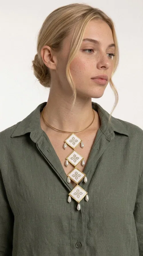, An embroidered statement necklace that instantly catches the eye — featuring geometric patterns inspired by Greek tradition and an aesthetic that speaks to both the past and the present.

Designed by Greek Jewelry Designer Aikaterini Chalkiadaki, this statement necklace combines white embroidered fabric with gold brass and white pearls in a composition reminiscent of folklore art with a contemporary touch. Four diamond-shaped embroidered elements are stacked vertically, one beneath the other, with teardrop pearls gracefully hanging from each corner. The piece is completed with a delicate gold rigid choker, giving it an airy yet striking presence.

Ideal for women who love long, eye-catching necklaces with a distinctive identity. It pairs perfectly with simple, solid-colored outfits — just as shown in the photo with the pink sleeveless top — or with boho looks for festivals and summer outings. It can also be combined with matching necklace and earring sets for a complete look.