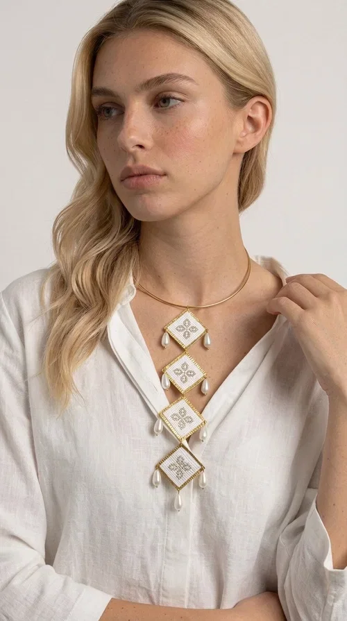 , An embroidered statement necklace that instantly catches the eye — featuring geometric patterns inspired by Greek tradition and an aesthetic that speaks to both the past and the present.

Designed by Greek Jewelry Designer Aikaterini Chalkiadaki, this statement necklace combines white embroidered fabric with gold brass and white pearls in a composition reminiscent of folklore art with a contemporary touch. Four diamond-shaped embroidered elements are stacked vertically, one beneath the other, with teardrop pearls gracefully hanging from each corner. The piece is completed with a delicate gold rigid choker, giving it an airy yet striking presence.

Ideal for women who love long, eye-catching necklaces with a distinctive identity. It pairs perfectly with simple, solid-colored outfits — just as shown in the photo with the pink sleeveless top — or with boho looks for festivals and summer outings. It can also be combined with matching necklace and earring sets for a complete look.