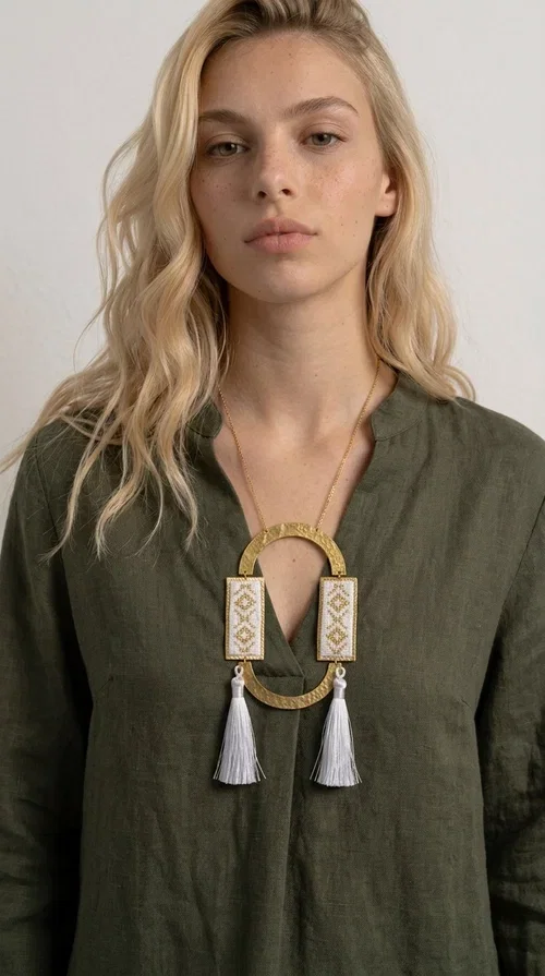 , An embroidered necklace that combines tradition with contemporary design — the Statement Necklace KOL-4757 is the ideal choice for every woman who wants to express herself through her jewelry. Designed by Greek Jewelry Designer Aikaterini Chalkiadaki, this handmade necklace is a unique combination of fabric and brass, inspired by folk art and the geometric motifs of Greek tradition.

Two embroidered rectangular plates with geometric patterns in white and gold frame a hammered gold brass circle, while three light-colored tassels add movement and character. The long gold chain completes a composition that is both statement-making and harmonious. Ideal for summer looks, casual outfits, and evening moments when you want to stand out.