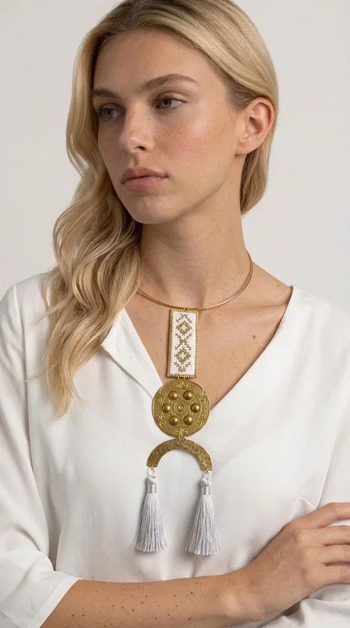 , A statement embroidered necklace that doesn't go unnoticed — featuring geometric patterns inspired by Greek tradition and an aesthetic that blends folk with contemporary style.

Designed by Greek Jewelry Designer Aikaterini Chalkiadaki, this handmade necklace consists of an embroidered rectangular section in white and gold with diamond-shaped geometric motifs, a round brass pendant with embossed details, and a semi-circular element from which two elegant white tassels hang. The piece is worn on a delicate gold torque-style chain that sits comfortably around the neck.

Ideal for those seeking striking jewelry that tells a story, this long necklace pairs equally well with a simple white shirt, a summer dress, or a boho outfit. Its white-and-gold palette makes it versatile for both day and evening wear.