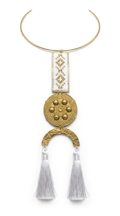 , A statement embroidered necklace that doesn't go unnoticed — featuring geometric patterns inspired by Greek tradition and an aesthetic that blends folk with contemporary style.

Designed by Greek Jewelry Designer Aikaterini Chalkiadaki, this handmade necklace consists of an embroidered rectangular section in white and gold with diamond-shaped geometric motifs, a round brass pendant with embossed details, and a semi-circular element from which two elegant white tassels hang. The piece is worn on a delicate gold torque-style chain that sits comfortably around the neck.

Ideal for those seeking striking jewelry that tells a story, this long necklace pairs equally well with a simple white shirt, a summer dress, or a boho outfit. Its white-and-gold palette makes it versatile for both day and evening wear.