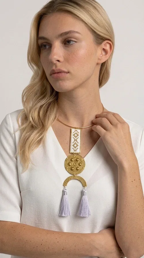 , A statement embroidered necklace that doesn't go unnoticed — featuring geometric patterns inspired by Greek tradition and an aesthetic that blends folk with contemporary style.

Designed by Greek Jewelry Designer Aikaterini Chalkiadaki, this handmade necklace consists of an embroidered rectangular section in white and gold with diamond-shaped geometric motifs, a round brass pendant with embossed details, and a semi-circular element from which two elegant white tassels hang. The piece is worn on a delicate gold torque-style chain that sits comfortably around the neck.

Ideal for those seeking striking jewelry that tells a story, this long necklace pairs equally well with a simple white shirt, a summer dress, or a boho outfit. Its white-and-gold palette makes it versatile for both day and evening wear.