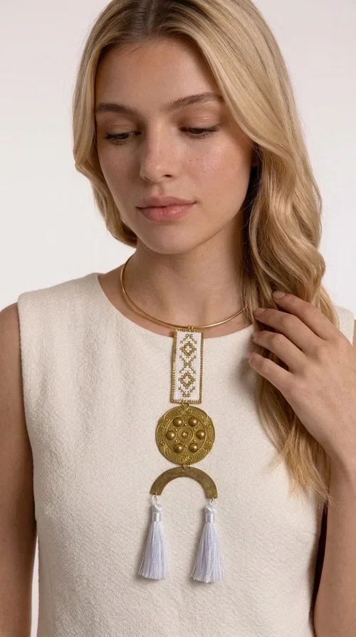 , A statement embroidered necklace that doesn't go unnoticed — featuring geometric patterns inspired by Greek tradition and an aesthetic that blends folk with contemporary style.

Designed by Greek Jewelry Designer Aikaterini Chalkiadaki, this handmade necklace consists of an embroidered rectangular section in white and gold with diamond-shaped geometric motifs, a round brass pendant with embossed details, and a semi-circular element from which two elegant white tassels hang. The piece is worn on a delicate gold torque-style chain that sits comfortably around the neck.

Ideal for those seeking striking jewelry that tells a story, this long necklace pairs equally well with a simple white shirt, a summer dress, or a boho outfit. Its white-and-gold palette makes it versatile for both day and evening wear.