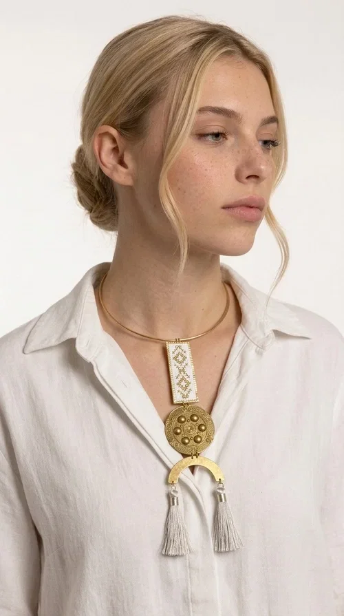 , A statement embroidered necklace that doesn't go unnoticed — featuring geometric patterns inspired by Greek tradition and an aesthetic that blends folk with contemporary style.

Designed by Greek Jewelry Designer Aikaterini Chalkiadaki, this handmade necklace consists of an embroidered rectangular section in white and gold with diamond-shaped geometric motifs, a round brass pendant with embossed details, and a semi-circular element from which two elegant white tassels hang. The piece is worn on a delicate gold torque-style chain that sits comfortably around the neck.

Ideal for those seeking striking jewelry that tells a story, this long necklace pairs equally well with a simple white shirt, a summer dress, or a boho outfit. Its white-and-gold palette makes it versatile for both day and evening wear.