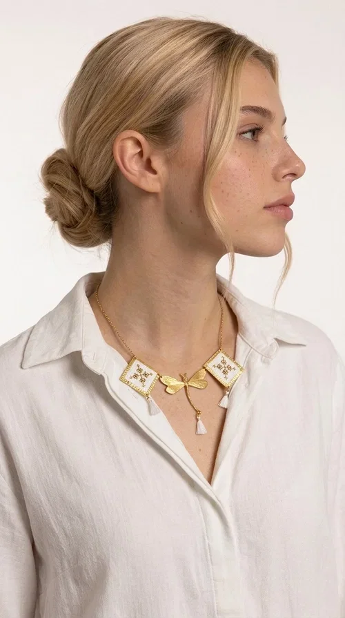 , The statement embroidered necklace in white with geometric patterns is a piece of jewelry that combines the traditional art of embroidery with a modern, striking result. Designed by Greek Jewelry Designer Aikaterini Chalkiadaki, this unique necklace showcases Greek craftsmanship in all its glory.

The necklace features two embroidered square elements in white with gold geometric motifs inspired by Greek folk tradition, joined by a gold dragonfly-shaped element. Three delicate white tassels add movement and joy to the piece, while the gold chain completes the look with elegance. The contrast between the white fabric and the brass creates a result that is both delicate and striking.

Ideal for summer looks, brunch, weddings, or evening outings, this handmade necklace pairs equally well with a simple white T-shirt or a boho dress. It also makes an excellent gift choice for women who love unique, handmade jewelry.