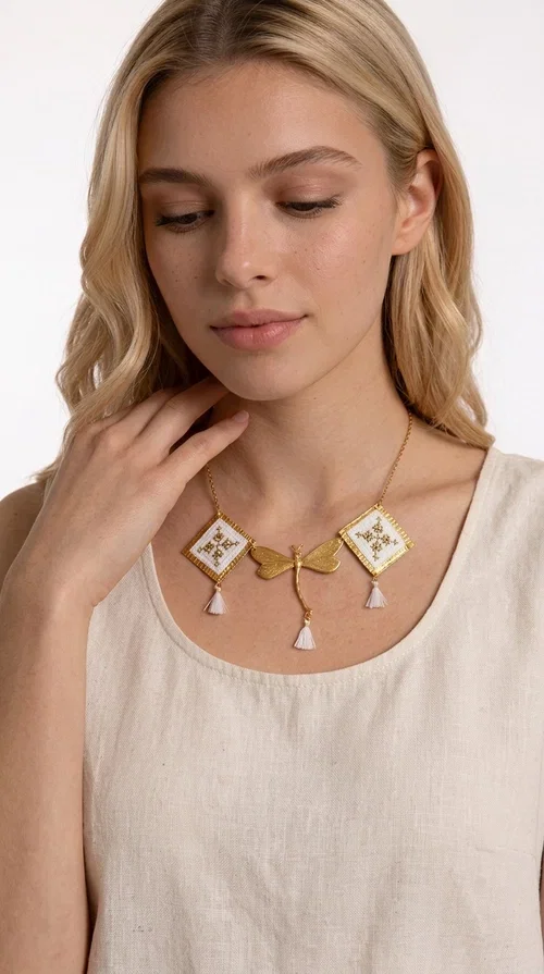 , The statement embroidered necklace in white with geometric patterns is a piece of jewelry that combines the traditional art of embroidery with a modern, striking result. Designed by Greek Jewelry Designer Aikaterini Chalkiadaki, this unique necklace showcases Greek craftsmanship in all its glory.

The necklace features two embroidered square elements in white with gold geometric motifs inspired by Greek folk tradition, joined by a gold dragonfly-shaped element. Three delicate white tassels add movement and joy to the piece, while the gold chain completes the look with elegance. The contrast between the white fabric and the brass creates a result that is both delicate and striking.

Ideal for summer looks, brunch, weddings, or evening outings, this handmade necklace pairs equally well with a simple white T-shirt or a boho dress. It also makes an excellent gift choice for women who love unique, handmade jewelry.