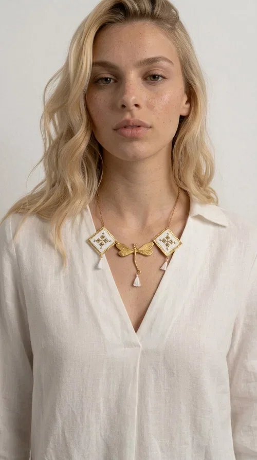 , The statement embroidered necklace in white with geometric patterns is a piece of jewelry that combines the traditional art of embroidery with a modern, striking result. Designed by Greek Jewelry Designer Aikaterini Chalkiadaki, this unique necklace showcases Greek craftsmanship in all its glory.

The necklace features two embroidered square elements in white with gold geometric motifs inspired by Greek folk tradition, joined by a gold dragonfly-shaped element. Three delicate white tassels add movement and joy to the piece, while the gold chain completes the look with elegance. The contrast between the white fabric and the brass creates a result that is both delicate and striking.

Ideal for summer looks, brunch, weddings, or evening outings, this handmade necklace pairs equally well with a simple white T-shirt or a boho dress. It also makes an excellent gift choice for women who love unique, handmade jewelry.