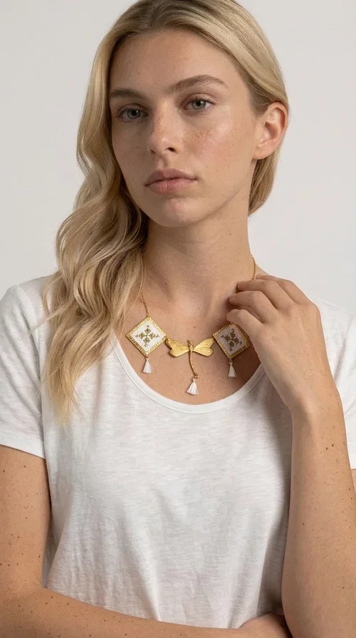 , The statement embroidered necklace in white with geometric patterns is a piece of jewelry that combines the traditional art of embroidery with a modern, striking result. Designed by Greek Jewelry Designer Aikaterini Chalkiadaki, this unique necklace showcases Greek craftsmanship in all its glory.

The necklace features two embroidered square elements in white with gold geometric motifs inspired by Greek folk tradition, joined by a gold dragonfly-shaped element. Three delicate white tassels add movement and joy to the piece, while the gold chain completes the look with elegance. The contrast between the white fabric and the brass creates a result that is both delicate and striking.

Ideal for summer looks, brunch, weddings, or evening outings, this handmade necklace pairs equally well with a simple white T-shirt or a boho dress. It also makes an excellent gift choice for women who love unique, handmade jewelry.