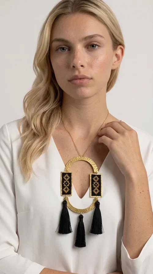 , A statement embroidered necklace that blends tradition with modern aesthetics — ideal for every woman who wants to express herself with style and character.

Designed by Greek Jewelry Designer Aikaterini Chalkiadaki, this striking long necklace is an ode to folk art and craftsmanship. Two rectangular embroidered plaques with geometric patterns in black and gold are framed by a golden brass arch, while three black tassels add movement and boho-chic charm. The gold-tone chain completes the composition with an elegant finishing touch.

This embroidered necklace pairs beautifully with simple white or ecru outfits that let the piece take center stage, as well as with evening looks that need that extra touch. Worn alone, it instantly turns any look into a statement.
