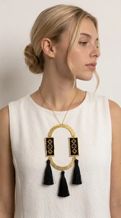 , A statement embroidered necklace that blends tradition with modern aesthetics — ideal for every woman who wants to express herself with style and character.

Designed by Greek Jewelry Designer Aikaterini Chalkiadaki, this striking long necklace is an ode to folk art and craftsmanship. Two rectangular embroidered plaques with geometric patterns in black and gold are framed by a golden brass arch, while three black tassels add movement and boho-chic charm. The gold-tone chain completes the composition with an elegant finishing touch.

This embroidered necklace pairs beautifully with simple white or ecru outfits that let the piece take center stage, as well as with evening looks that need that extra touch. Worn alone, it instantly turns any look into a statement.