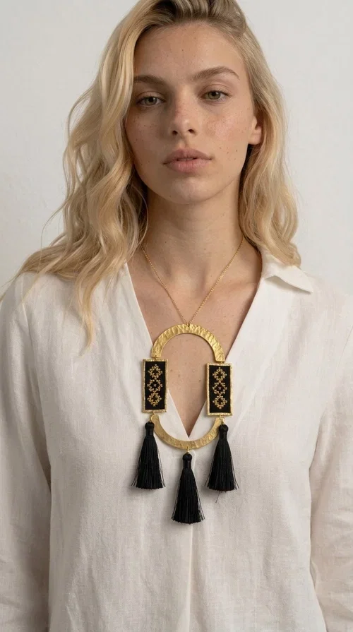 , A statement embroidered necklace that blends tradition with modern aesthetics — ideal for every woman who wants to express herself with style and character.

Designed by Greek Jewelry Designer Aikaterini Chalkiadaki, this striking long necklace is an ode to folk art and craftsmanship. Two rectangular embroidered plaques with geometric patterns in black and gold are framed by a golden brass arch, while three black tassels add movement and boho-chic charm. The gold-tone chain completes the composition with an elegant finishing touch.

This embroidered necklace pairs beautifully with simple white or ecru outfits that let the piece take center stage, as well as with evening looks that need that extra touch. Worn alone, it instantly turns any look into a statement.
