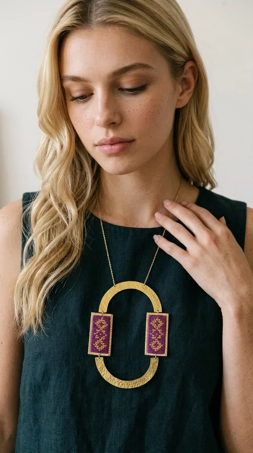 , An embroidered necklace that instantly catches the eye — the Statement Necklace KOL-4753 combines the tradition of Greek embroidery art with a modern, geometric aesthetic. Designed by Greek Jewelry Designer Aikaterini Chalkiadaki, this striking long necklace is the ultimate statement piece for every woman who appreciates handmade artistry and uniqueness.

The necklace features two rectangular fabric elements in deep burgundy, adorned with gold embroidery of geometric patterns inspired by folklore aesthetics. The embroidered sections are framed by shiny gold-toned brass, forming a large oval shape — a bold design choice that highlights the uniqueness of the piece. The gold-toned chain offers an elegant long line that pairs perfectly with simple, monochrome outfits.

Ideal for stylish daytime looks as well as evening outings, this necklace suits both casual and formal ensembles equally well. It gives your look an authentic, artistic character.