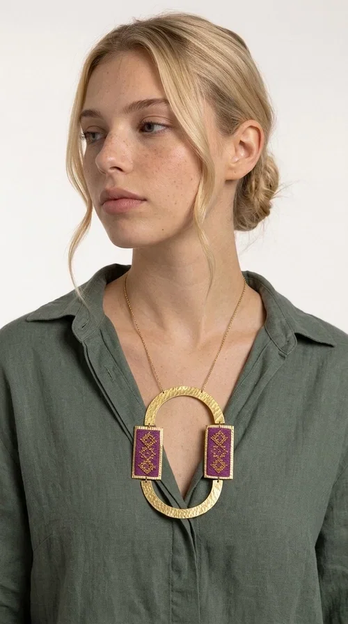 , An embroidered necklace that instantly catches the eye — the Statement Necklace KOL-4753 combines the tradition of Greek embroidery art with a modern, geometric aesthetic. Designed by Greek Jewelry Designer Aikaterini Chalkiadaki, this striking long necklace is the ultimate statement piece for every woman who appreciates handmade artistry and uniqueness.

The necklace features two rectangular fabric elements in deep burgundy, adorned with gold embroidery of geometric patterns inspired by folklore aesthetics. The embroidered sections are framed by shiny gold-toned brass, forming a large oval shape — a bold design choice that highlights the uniqueness of the piece. The gold-toned chain offers an elegant long line that pairs perfectly with simple, monochrome outfits.

Ideal for stylish daytime looks as well as evening outings, this necklace suits both casual and formal ensembles equally well. It gives your look an authentic, artistic character.