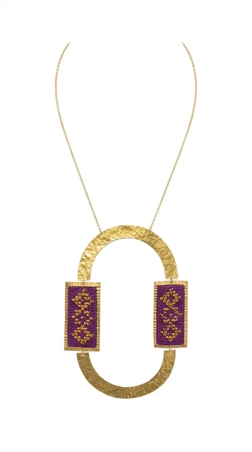 , An embroidered necklace that instantly catches the eye — the Statement Necklace KOL-4753 combines the tradition of Greek embroidery art with a modern, geometric aesthetic. Designed by Greek Jewelry Designer Aikaterini Chalkiadaki, this striking long necklace is the ultimate statement piece for every woman who appreciates handmade artistry and uniqueness.

The necklace features two rectangular fabric elements in deep burgundy, adorned with gold embroidery of geometric patterns inspired by folklore aesthetics. The embroidered sections are framed by shiny gold-toned brass, forming a large oval shape — a bold design choice that highlights the uniqueness of the piece. The gold-toned chain offers an elegant long line that pairs perfectly with simple, monochrome outfits.

Ideal for stylish daytime looks as well as evening outings, this necklace suits both casual and formal ensembles equally well. It gives your look an authentic, artistic character.