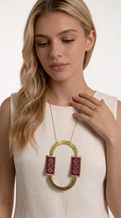 , An embroidered necklace that instantly catches the eye — the Statement Necklace KOL-4753 combines the tradition of Greek embroidery art with a modern, geometric aesthetic. Designed by Greek Jewelry Designer Aikaterini Chalkiadaki, this striking long necklace is the ultimate statement piece for every woman who appreciates handmade artistry and uniqueness.

The necklace features two rectangular fabric elements in deep burgundy, adorned with gold embroidery of geometric patterns inspired by folklore aesthetics. The embroidered sections are framed by shiny gold-toned brass, forming a large oval shape — a bold design choice that highlights the uniqueness of the piece. The gold-toned chain offers an elegant long line that pairs perfectly with simple, monochrome outfits.

Ideal for stylish daytime looks as well as evening outings, this necklace suits both casual and formal ensembles equally well. It gives your look an authentic, artistic character.