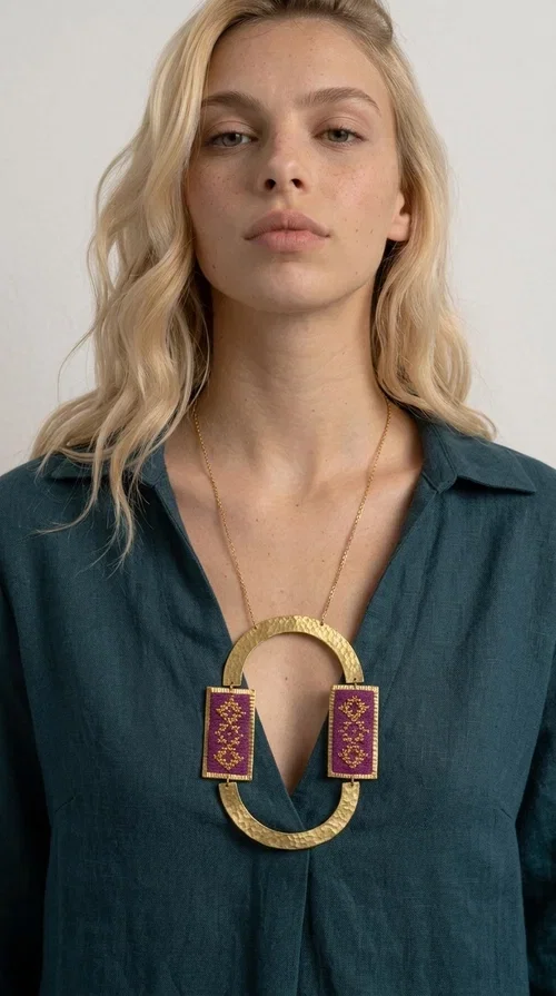 , An embroidered necklace that instantly catches the eye — the Statement Necklace KOL-4753 combines the tradition of Greek embroidery art with a modern, geometric aesthetic. Designed by Greek Jewelry Designer Aikaterini Chalkiadaki, this striking long necklace is the ultimate statement piece for every woman who appreciates handmade artistry and uniqueness.

The necklace features two rectangular fabric elements in deep burgundy, adorned with gold embroidery of geometric patterns inspired by folklore aesthetics. The embroidered sections are framed by shiny gold-toned brass, forming a large oval shape — a bold design choice that highlights the uniqueness of the piece. The gold-toned chain offers an elegant long line that pairs perfectly with simple, monochrome outfits.

Ideal for stylish daytime looks as well as evening outings, this necklace suits both casual and formal ensembles equally well. It gives your look an authentic, artistic character.