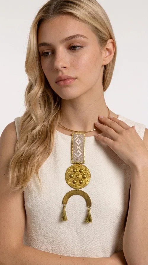, The ecru embroidered statement necklace with geometric patterns is a piece of jewelry that combines the traditional art of embroidery with modern aesthetics, creating an immediately eye-catching result. Designed by Greek jewelry designer Aikaterini Chalkiadaki, this handmade necklace is a distinctive choice for women who appreciate uniqueness and authentic craftsmanship.

The necklace features a rectangular embroidered section with fine geometric motifs in an ecru shade, paired with a circular brass element featuring embossed details and a semicircular arch. Three dangling khaki-olive tassels add movement and a boho-folklore character, while the base of the necklace consists of a semi-rigid metallic neckwire that rests comfortably on the collarbone. The combination of brass and fabric makes it lightweight to wear, despite its striking appearance.

Ideal for summer outfits, boho looks, ethnic, and festival styles, it pairs perfectly with linen shirts, maxi dresses, or simple tops. It is also an excellent gift idea for women who love large, statement jewelry.