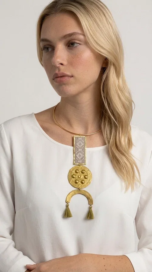 , The ecru embroidered statement necklace with geometric patterns is a piece of jewelry that combines the traditional art of embroidery with modern aesthetics, creating an immediately eye-catching result. Designed by Greek jewelry designer Aikaterini Chalkiadaki, this handmade necklace is a distinctive choice for women who appreciate uniqueness and authentic craftsmanship.

The necklace features a rectangular embroidered section with fine geometric motifs in an ecru shade, paired with a circular brass element featuring embossed details and a semicircular arch. Three dangling khaki-olive tassels add movement and a boho-folklore character, while the base of the necklace consists of a semi-rigid metallic neckwire that rests comfortably on the collarbone. The combination of brass and fabric makes it lightweight to wear, despite its striking appearance.

Ideal for summer outfits, boho looks, ethnic, and festival styles, it pairs perfectly with linen shirts, maxi dresses, or simple tops. It is also an excellent gift idea for women who love large, statement jewelry.