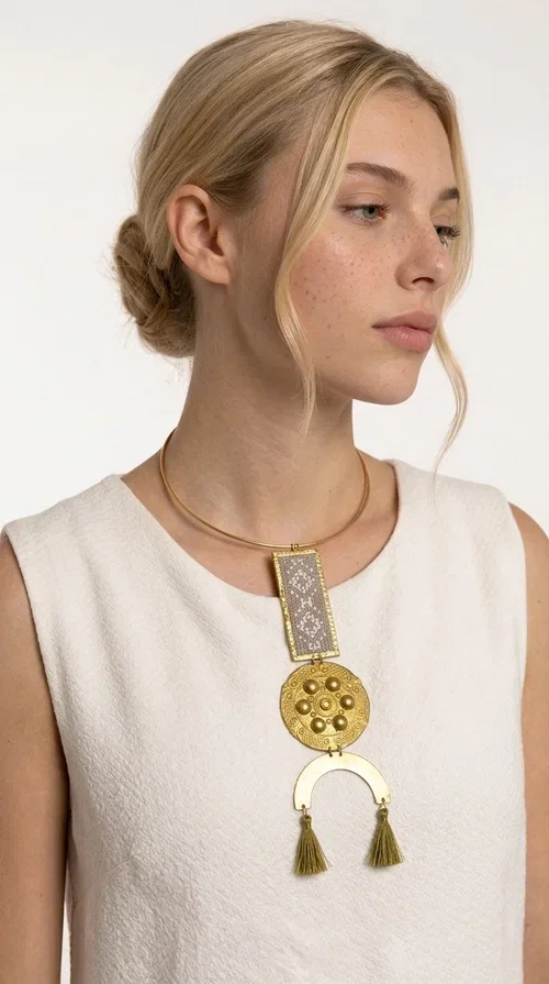 , The ecru embroidered statement necklace with geometric patterns is a piece of jewelry that combines the traditional art of embroidery with modern aesthetics, creating an immediately eye-catching result. Designed by Greek jewelry designer Aikaterini Chalkiadaki, this handmade necklace is a distinctive choice for women who appreciate uniqueness and authentic craftsmanship.

The necklace features a rectangular embroidered section with fine geometric motifs in an ecru shade, paired with a circular brass element featuring embossed details and a semicircular arch. Three dangling khaki-olive tassels add movement and a boho-folklore character, while the base of the necklace consists of a semi-rigid metallic neckwire that rests comfortably on the collarbone. The combination of brass and fabric makes it lightweight to wear, despite its striking appearance.

Ideal for summer outfits, boho looks, ethnic, and festival styles, it pairs perfectly with linen shirts, maxi dresses, or simple tops. It is also an excellent gift idea for women who love large, statement jewelry.