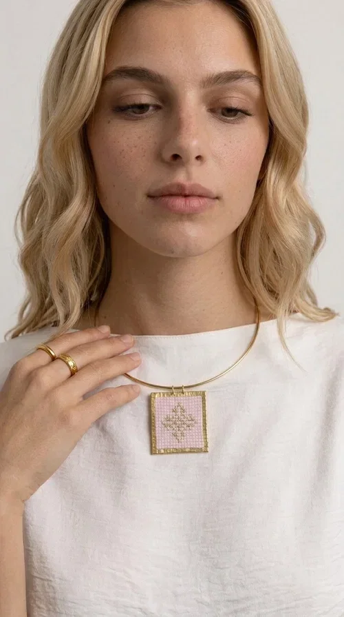 , An embroidered necklace that combines the traditional art of embroidery with modern design, creating a piece of jewelry that stands out instantly.

Designed by Greek Jewelry Designer Aikaterini Chalkiadaki, this statement necklace features a rectangular embroidered motif in soft pink tones with gold geometric details inspired by Greek folk tradition. The gold brass frame highlights the embroidered fabric, while the delicate semicircular chain gives the whole piece an airy and elegant character.

Ideal for women who love striking jewelry with soul, this handmade necklace pairs perfectly with both casual and more formal outfits — just as shown here over a classic beige jacket. Bring folk aesthetics into your everyday look in a completely modern way.