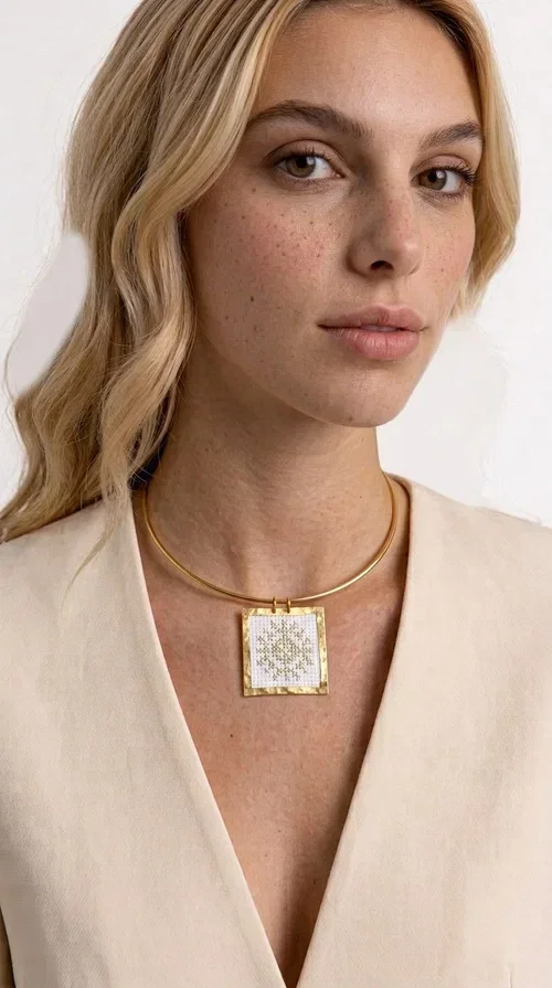 , An embroidered necklace that combines tradition with modern aesthetics — the Statement Necklace KOL-4721 is a handmade creation that stands out for its refined detail and unique character. Designed by Greek Jewelry Designer Aikaterini Chalkiadaki, this piece pays tribute to the folk Greek art of embroidery, rendering geometric motifs on white fabric with gold details.

The rectangular embroidered pendant, featuring a handcrafted gold-tone brass frame, hangs from a classic gold omega-style necklace chain, which adds stability and elegance to the overall look. The folklore geometric patterns on the white fabric create an eye-catching contrast, while the overall aesthetic moves between ethnic and contemporary style.

Ideal for pairing with linen or wool clothing in neutral tones, it suits both casual and more formal looks. A true statement piece for the neck that needs no other additions to shine.